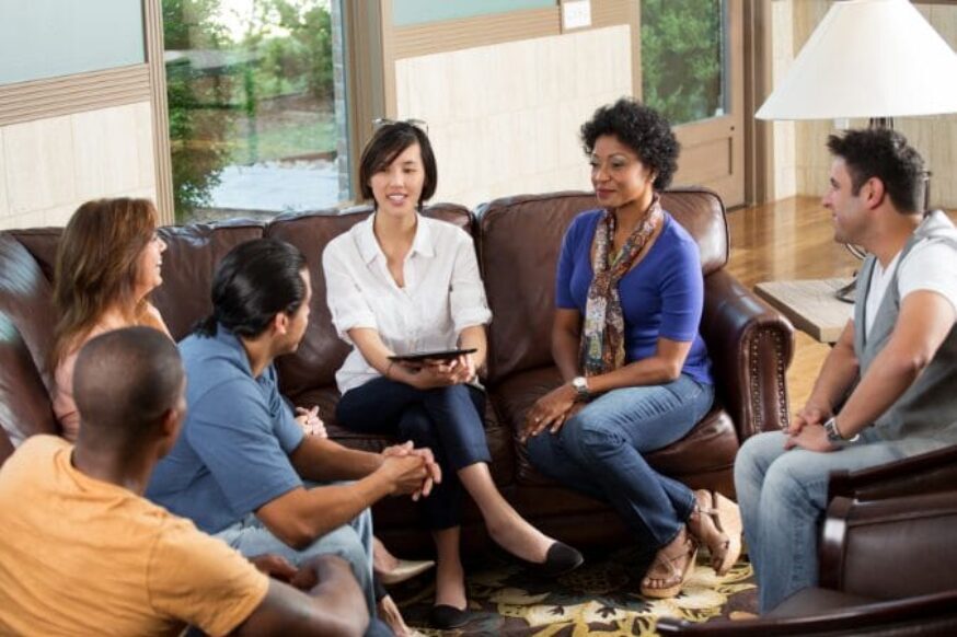 Five adults are having a conversation in a living room setting with a sofa and armchair.