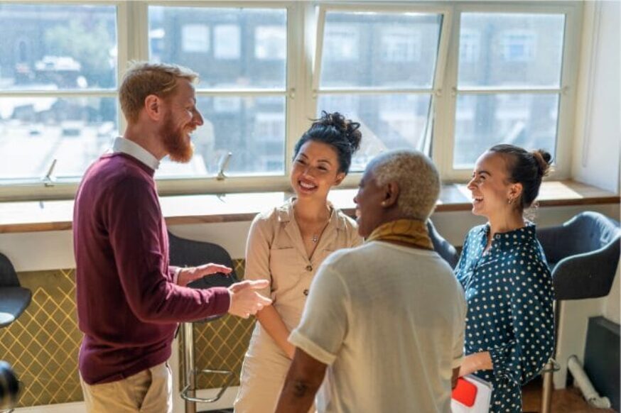 Four people appear to be having a conversation in a bright, indoor setting with large windows.