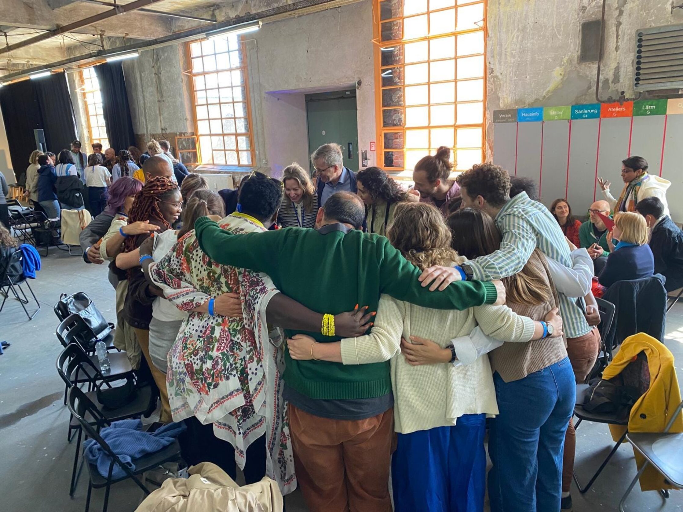 A wide shot shows a large, diverse group of people gathered indoors in what appears to be a spacious, industrial-style room with high ceilings and large windows providing natural light. The focus is on a central huddle of approximately ten individuals standing in a close circle, embracing each other with their arms around shoulders and backs. They seem to be of various ages and likely diverse ethnic backgrounds. Some are smiling and appear connected.