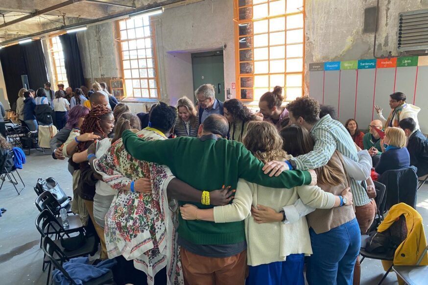 A wide shot shows a large, diverse group of people gathered indoors in what appears to be a spacious, industrial-style room with high ceilings and large windows providing natural light. The focus is on a central huddle of approximately ten individuals standing in a close circle, embracing each other with their arms around shoulders and backs. They seem to be of various ages and likely diverse ethnic backgrounds. Some are smiling and appear connected.
