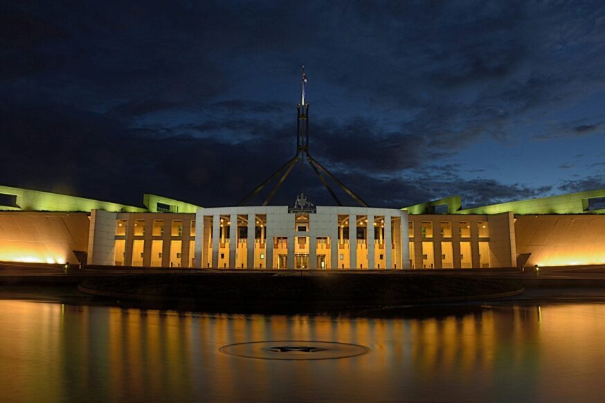 A nighttime, long shot captures the illuminated Parliament House in Canberra, Australia, reflected in the still waters of a foreground pool. The modern, white building features a central flagpole structure reaching towards the dark, cloudy sky. Warm, golden light emanates from the building's many windows and architectural details, contrasting with the cool tones of the night sky and the water's surface. The reflection in the water creates a symmetrical and slightly distorted image of the building and its lights.