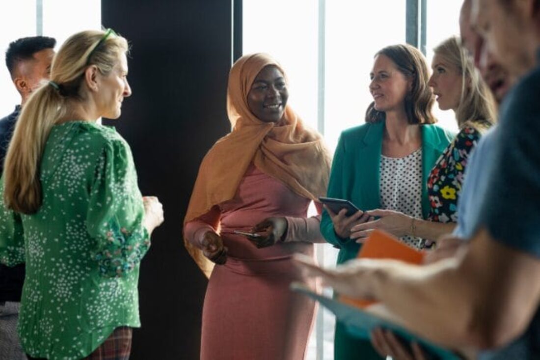 A group of diverse people gathered in a bright room, engaging in conversation. A woman in a green polka-dot blouse talks with a smiling Black woman in a peach hijab and pink dress, who holds a card. Two other women look at a phone to their right.