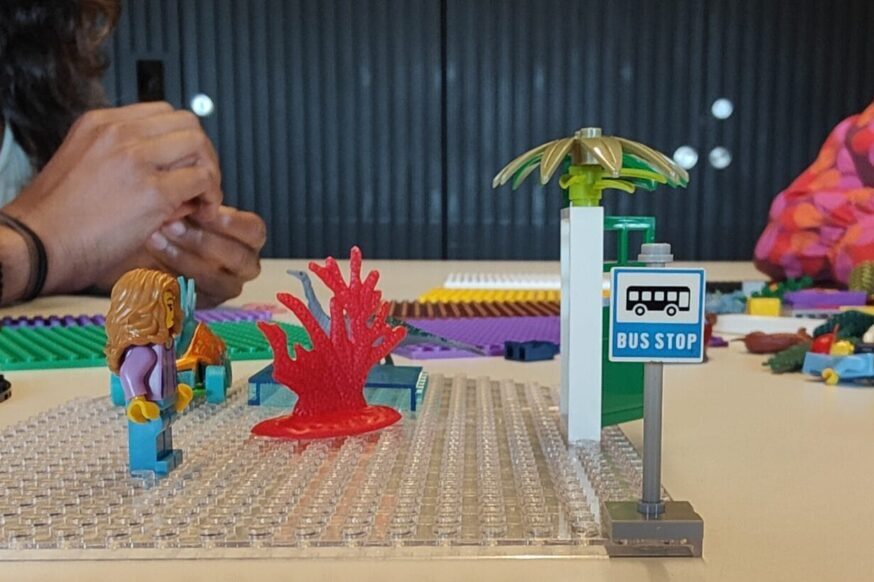 A medium shot of a table with several people building with LEGOs. In the center, a translucent grey baseplate has a bus stop with a small palm tree, a red coral piece, and a LEGO figurine. A person wearing a colorful patterned shirt is building in the background on the right. Another person is building in the background on the left, wearing a grey sweater.