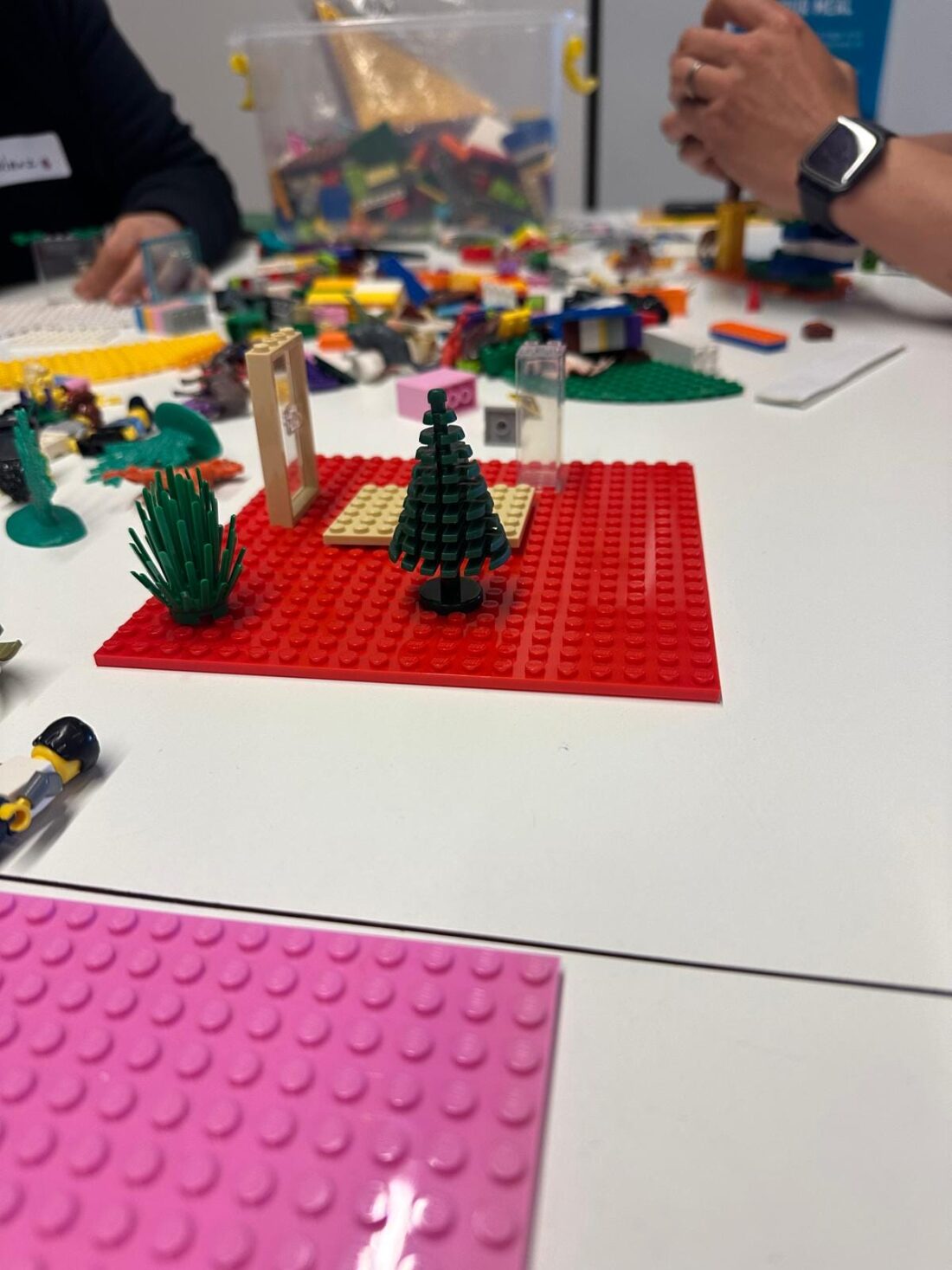 A close-up, high-angle shot of a white table covered with various LEGO creations and pieces. In the foreground, there is a large, red LEGO baseplate with a small house-like structure and two green LEGO plants built on it. To the left, a pink LEGO baseplate is partially visible. In the background, other participants' hands and more LEGO pieces are scattered on the table.