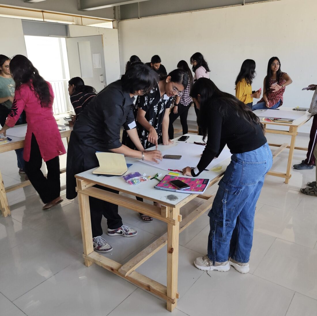 An indoor photograph of a group of young people, primarily women, working together around light wood tables in a brightly lit, spacious room. Two people in the center foreground, one wearing a black shirt and the other a black sweater and blue jeans, are bent over a large sheet of paper on a table, seemingly collaborating or instructing. Other participants are scattered throughout the background, also engaged in similar activities at other tables. The room has a white tile floor, white walls, and an open doorway on the left leading to the outside.