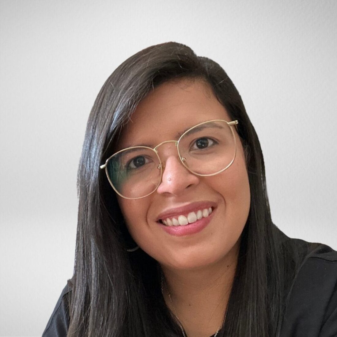 A headshot of a smiling young woman with long, dark hair, wearing a black shirt and thin, gold-rimmed glasses. The background is a plain, light grey/white.