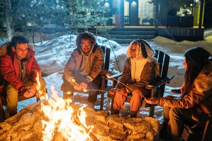 An outdoor, nighttime photo shows four young adults sitting in wooden Adirondack chairs around a roaring bonfire in a snowy or frost-covered area. They are all bundled up in winter jackets and appear to be roasting marshmallows on sticks. They are laughing and smiling, enjoying the fire and each other's company. In the background, there is a modern-looking wooden building, possibly a cabin or lodge, partially visible.