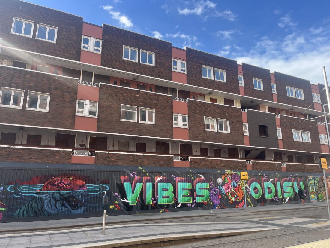 A large, multi-story residential building with brown and reddish-pink facades is visible under a blue sky with white clouds. In the foreground, there is a low metal barrier or hoarding along the sidewalk and tram tracks, which is completely covered with vibrant, colorful graffiti art. The graffiti features a large green word 