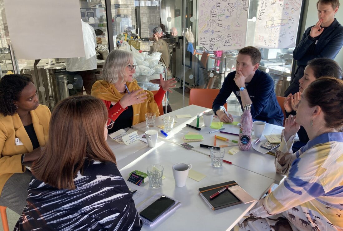 A group of eight people is gathered around a square white table in a brightly lit room for a discussion or meeting. Six people are seated, and two men are standing behind the table. One woman in the center, wearing a yellow wrap over a red top, is speaking animatedly with her hands. Various notebooks, pens, markers, coffee cups, and a pink water bottle are scattered on the table. In the background, there is a large whiteboard covered with diagrams and writing, and a glass partition reveals a kitchen or catering area.