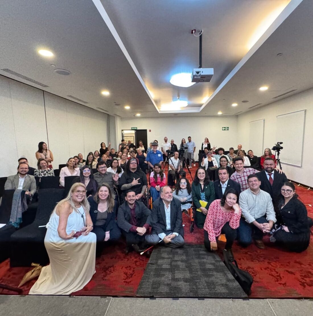 A wide group photo of a diverse audience posing together inside a modern conference room with red carpeting, with some individuals sitting in rows of chairs and a few people kneeling or sitting on the floor in the foreground.