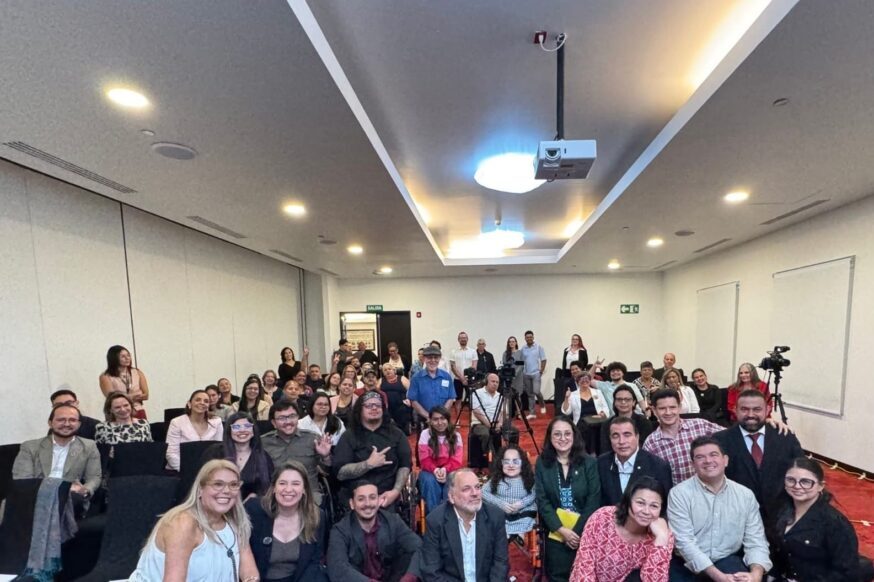 A wide group photo of a diverse audience posing together inside a modern conference room with red carpeting, with some individuals sitting in rows of chairs and a few people kneeling or sitting on the floor in the foreground.