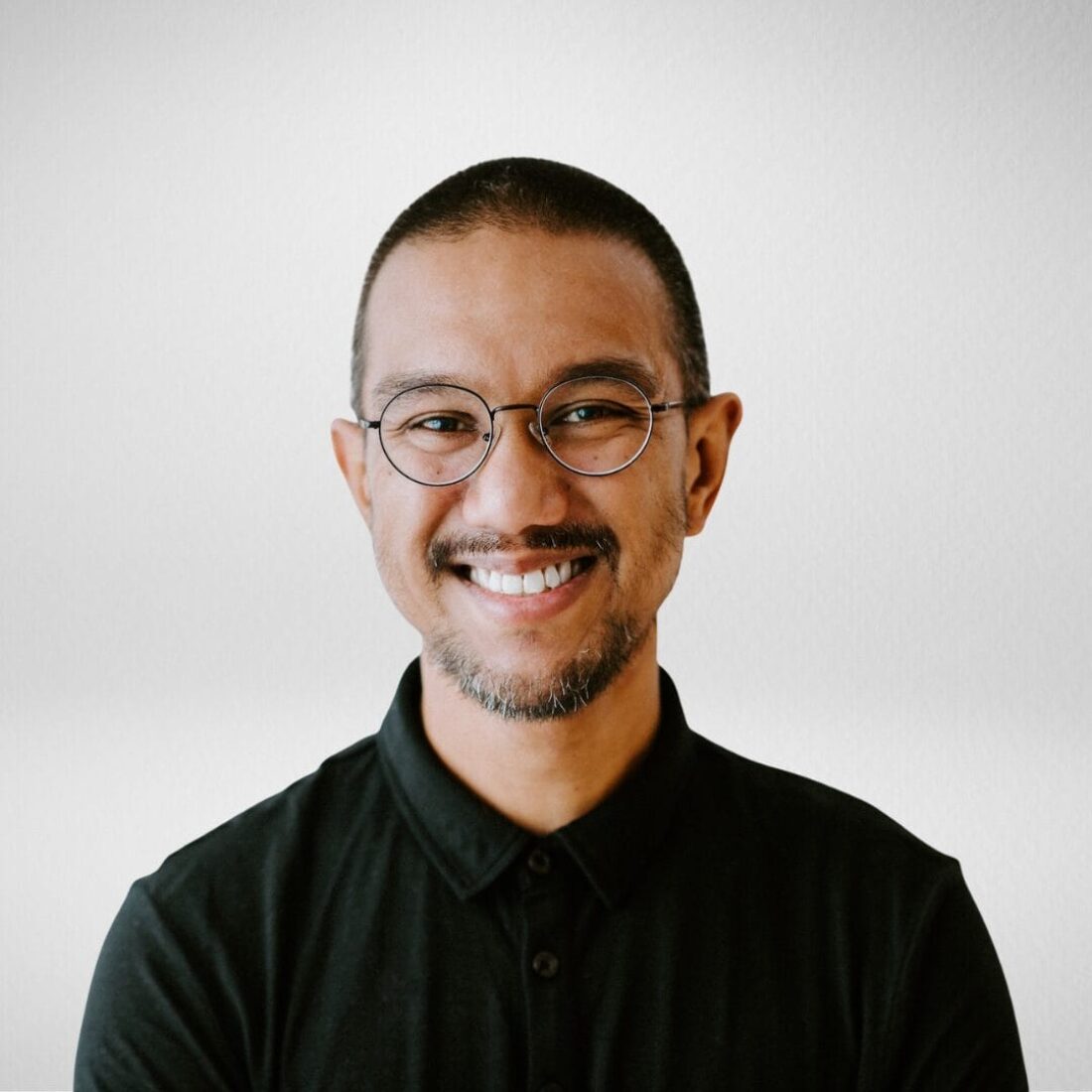 A smiling man with a shaved head, short beard, and round, thin-framed glasses, wearing a black collared shirt against a plain, light background.