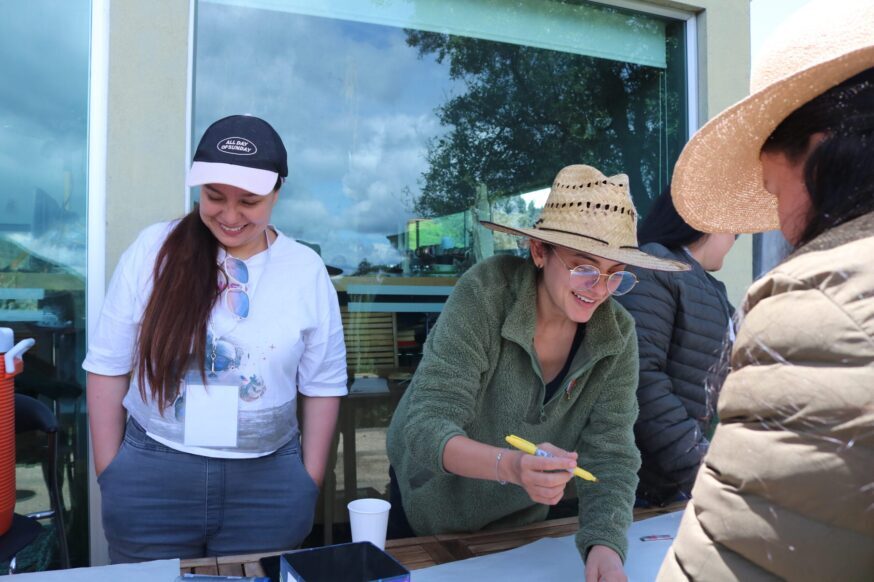 A smiling woman wearing a straw hat and green fleece sweater writes on a large sheet of paper with a yellow marker at an outdoor table. Next to her, a smiling woman in a white t-shirt, baseball cap, and gray jeans looks on with her hands in her pockets.