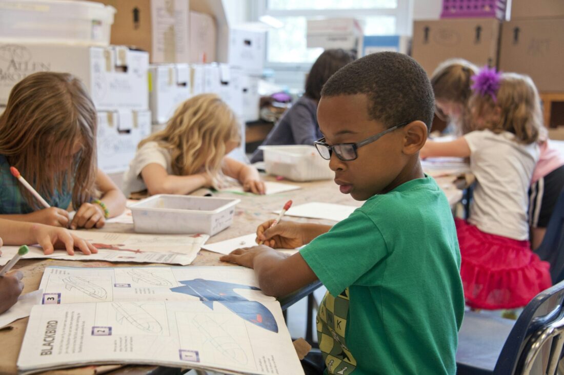A young boy with glasses in a green t-shirt writes on paper while sitting at a table surrounded by other school children in a classroom.