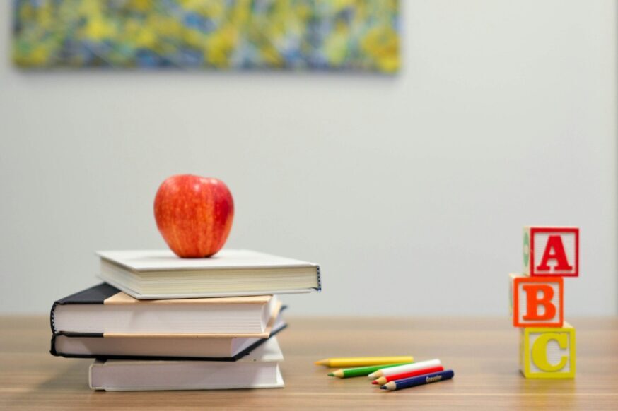 A stack of books topped with a red apple, scattered colored pencils, and toy letter blocks spelling 
