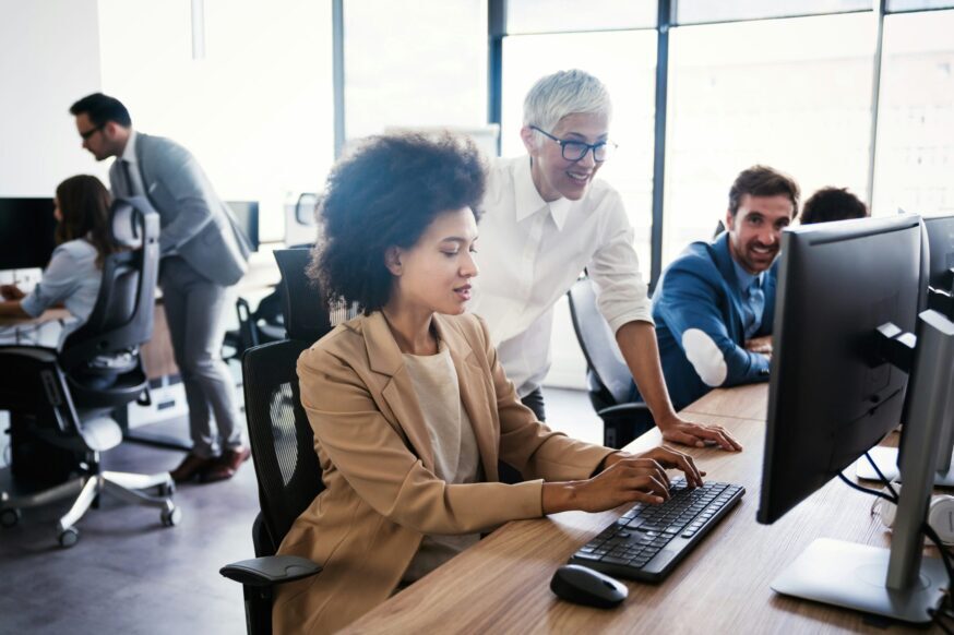 A professional office scene where a woman with curly dark hair sits at a long wooden desk typing on a computer keyboard. A senior woman with short grey hair and glasses leans over her shoulder, smiling as they look at the desktop screen together. In the background, other colleagues are seated at desks or standing in a modern, brightly lit open-plan workspace with large windows.