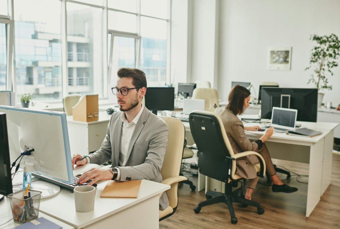 A man wearing glasses and a grey suit blazer working at a computer in a modern, open-plan office, with a colleague working in the background.