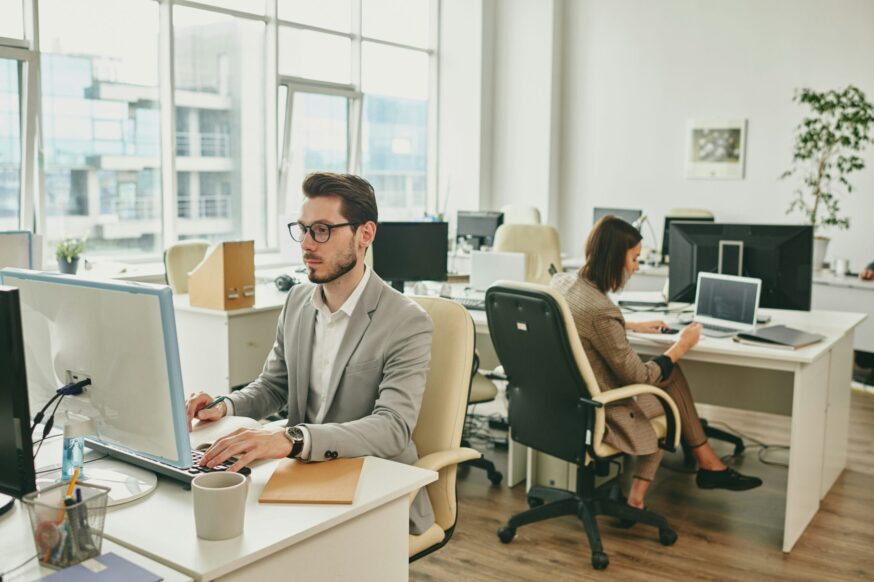 A man wearing glasses and a grey suit blazer working at a computer in a modern, open-plan office, with a colleague working in the background.